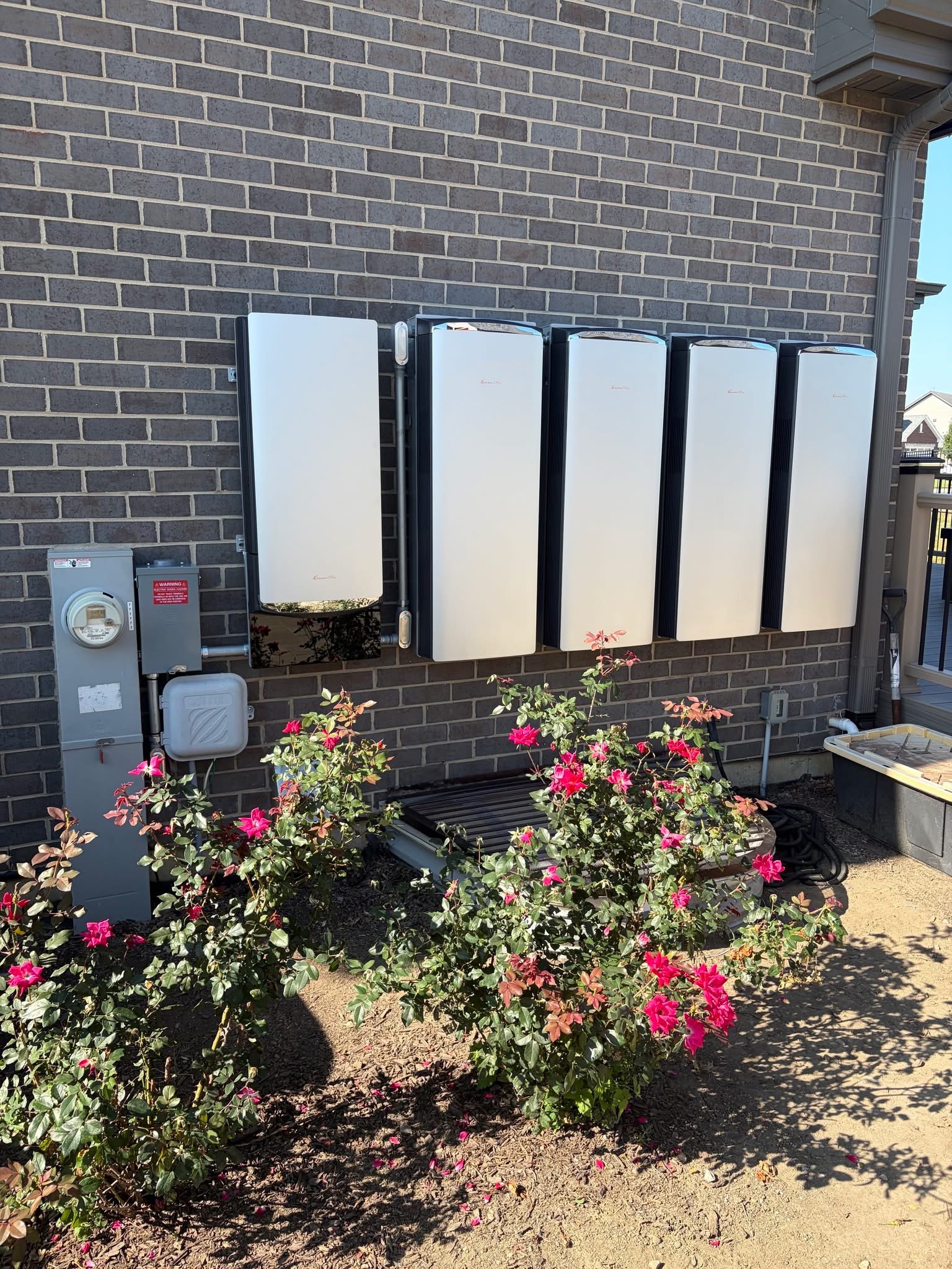 Home battery backup system with five wall-mounted units installed on a brick home exterior, above a bed of roses
