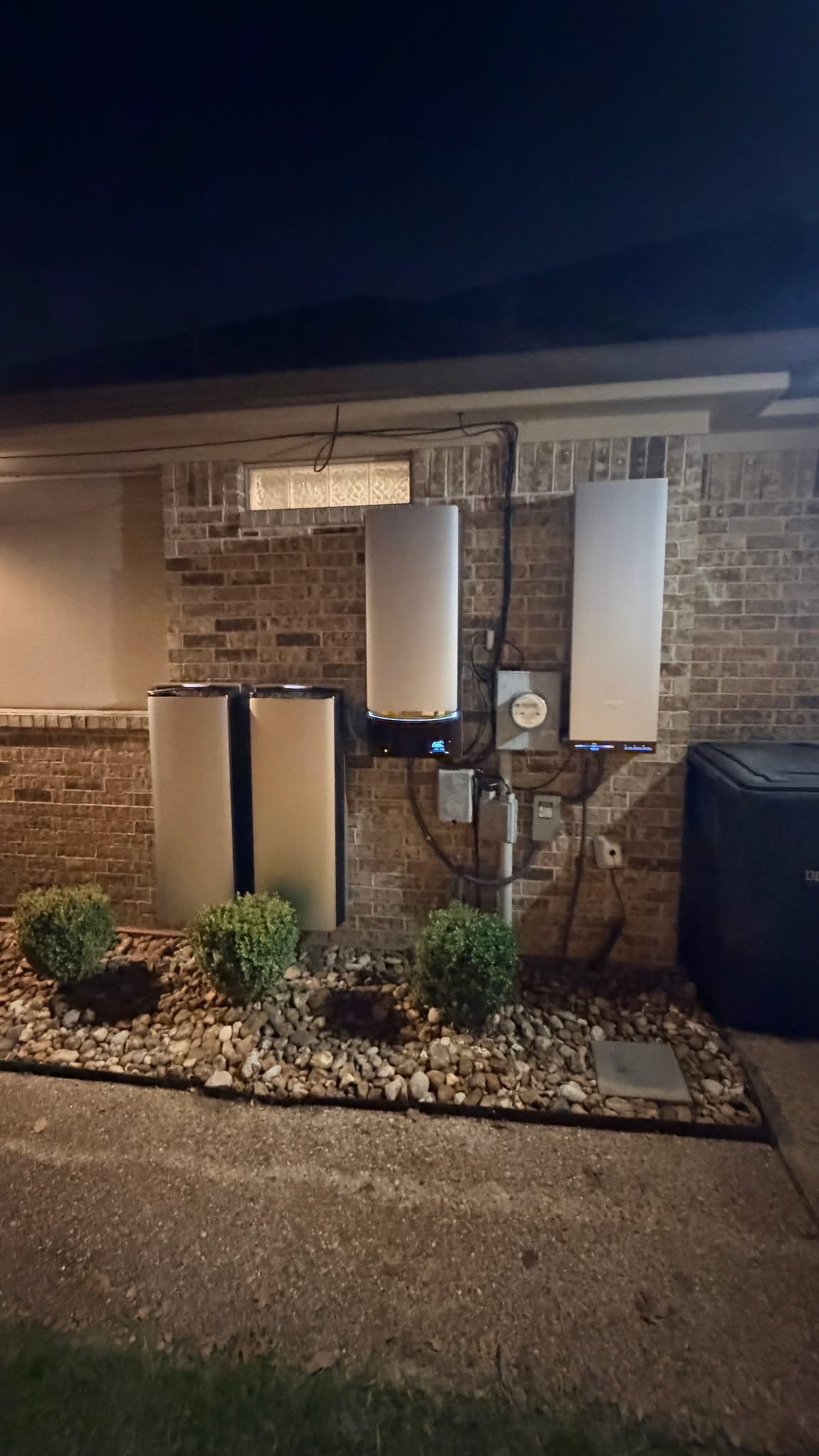 Home battery backup system installed on a brick home exterior at night, above a landscaped rock bed